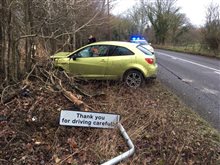A crashed yellow car next to a Thank you for driving carefully village sign with a police car behind