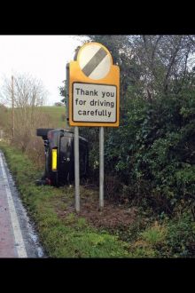 A car overturned on its side next to a Thank you for driving carefully road sign on a country lane